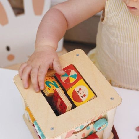 A toddler in a light pink dress interacts with the wooden activity cube by Mentari, reaching out to spin the patterned panels on the side.