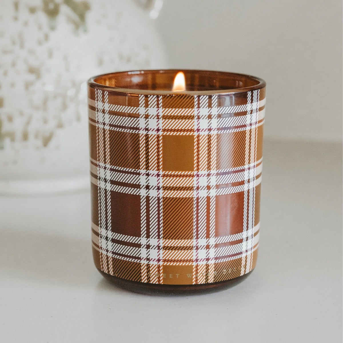 A lit candle in a brown glass jar with a white and brown plaid pattern, set against a softly blurred light background.
