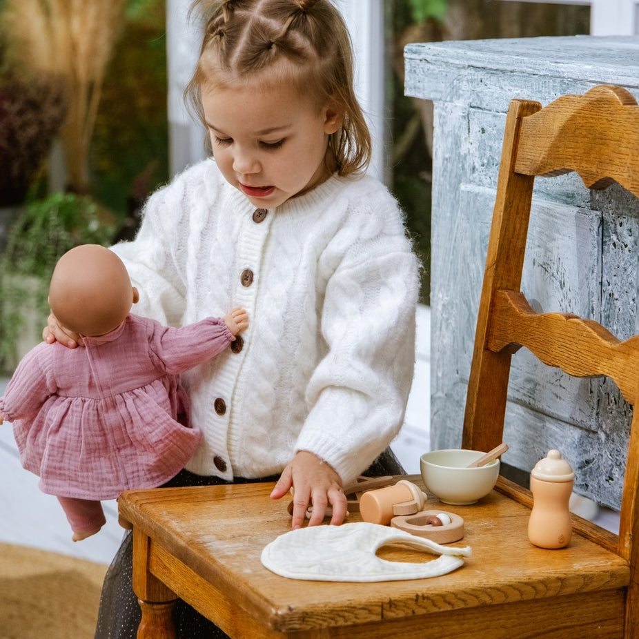 A young girl with braided hair sits on a wooden chair, wearing a white cable-knit cardigan and playing with a pink doll. In front of her on the wooden table are various toy props, including a white bib, a small wooden bottle, and other play items, set against a rustic indoor background with a window and a weathered blue-gray cabinet.