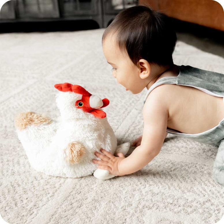 A baby lying on a textured rug, reaching out to touch a fluffy white Warmies chicken plush toy with a red comb and wattle.