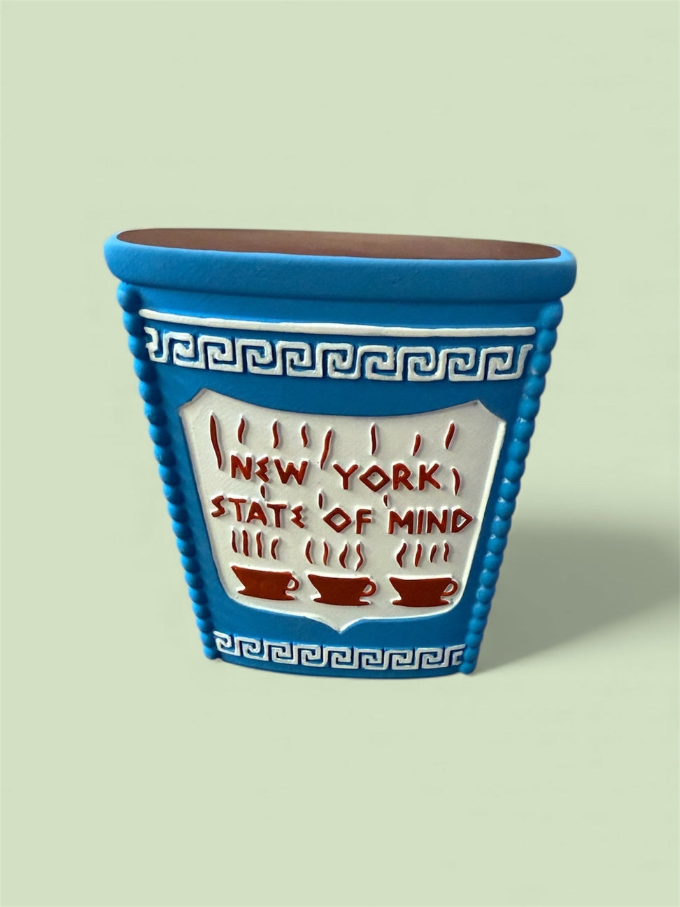 A blue ceramic coffee mug with a Greek key pattern and a white center panel that reads 'NEW YORK STATE OF MIND' with red coffee cups and steam, shown on a pale green background.