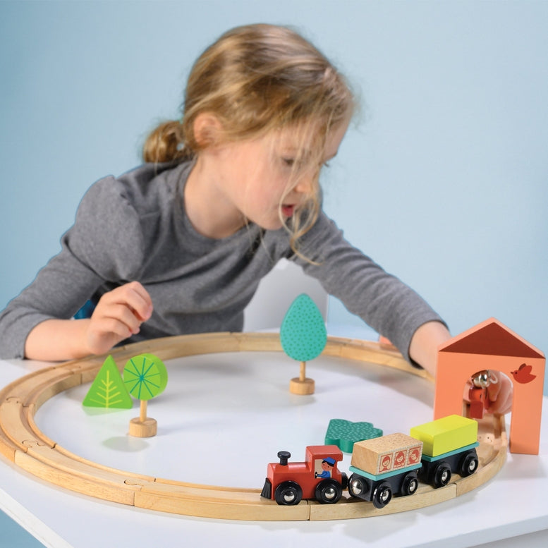 A young child playing with the My First Wooden Train Set by Mentari, assembling the wooden tracks and setting up a toy train, house, and trees on a white surface.