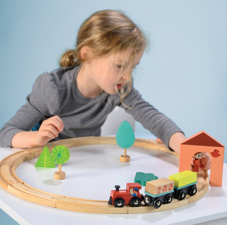 A young child playing with the My First Wooden Train Set by Mentari, assembling the wooden tracks and setting up a toy train, house, and trees on a white surface.