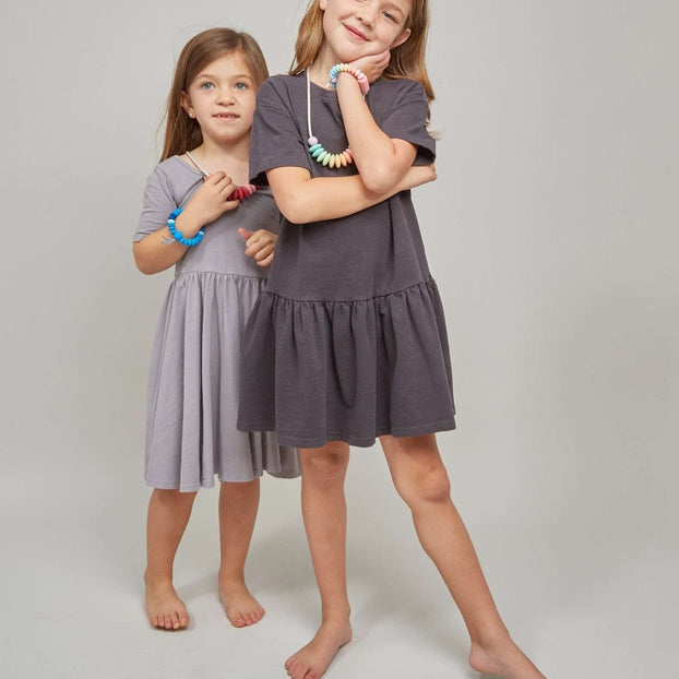 Two young girls modeling short-sleeve tiered dresses in a studio setting. The girl in the foreground wears a dark charcoal gray dress and poses with her arms crossed, while the girl behind her wears a light gray dress.