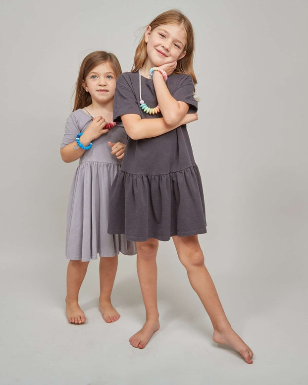 Two young girls modeling short-sleeve tiered dresses in a studio setting. The girl in the foreground wears a dark charcoal gray dress and poses with her arms crossed, while the girl behind her wears a light gray dress.
