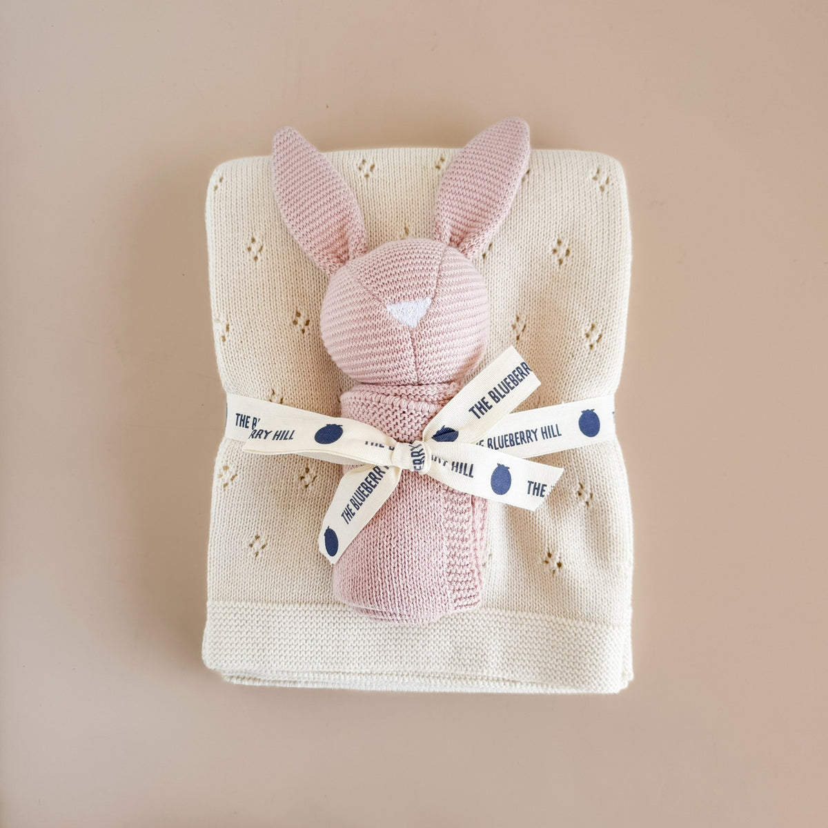 A folded cream-colored knit baby blanket with small eyelet details, featuring a knitted pink bunny plush tucked inside and tied with a cream ribbon printed with navy blue dots and the text 'THE BLUEBERRY HILL'.