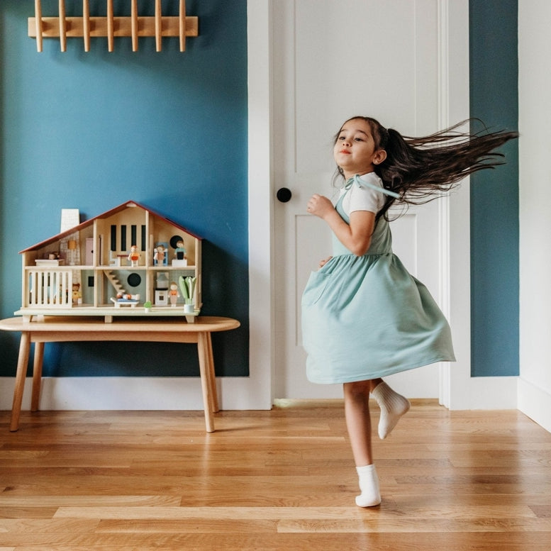 A young child in a light-colored dress skips playfully across a hardwood floor in a brightly lit room, with the Wooden Cedar Chalet Modernist Dollhouse by Mentari resting on a low wooden bench against a deep teal accent wall.