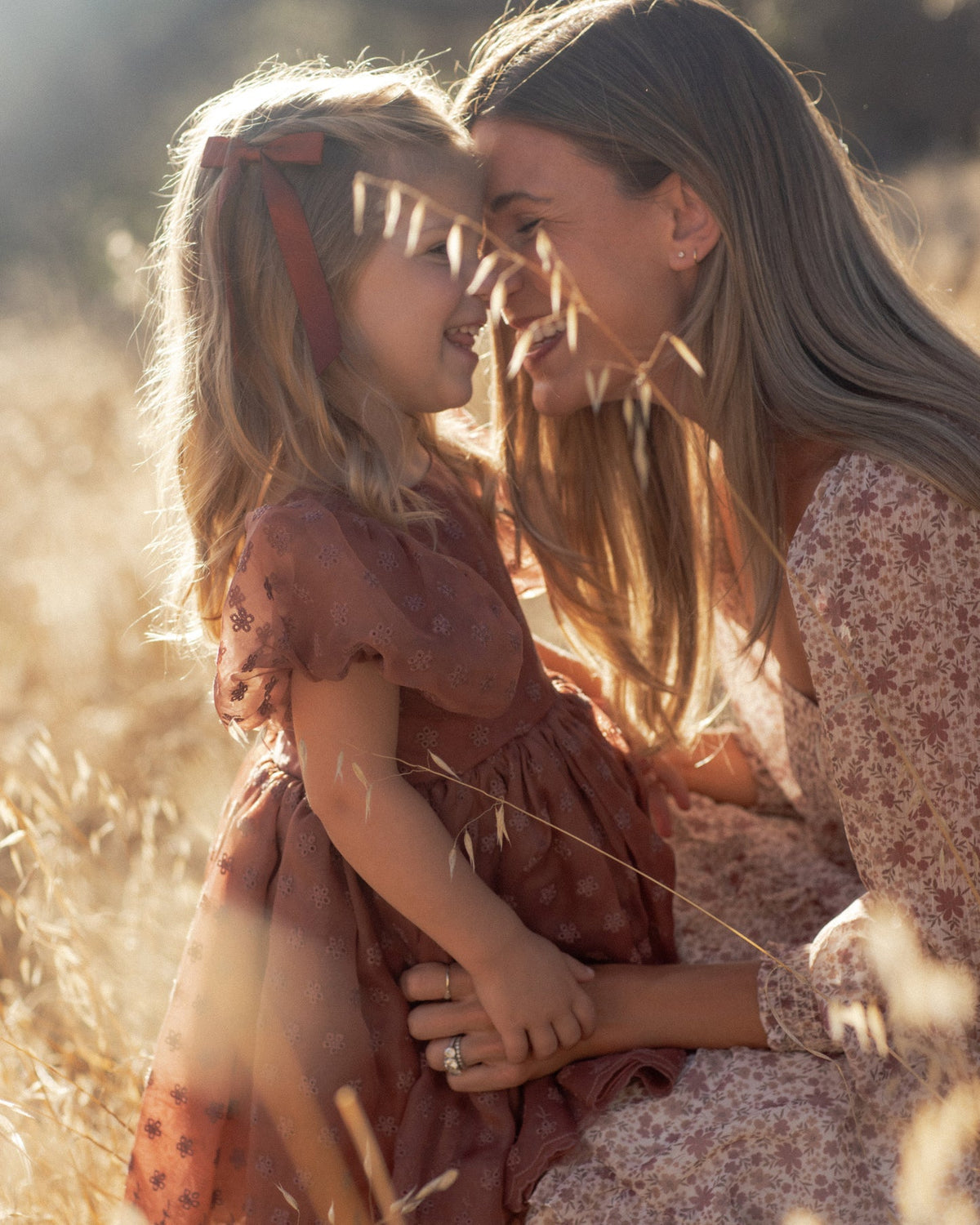 A woman and a young girl in a sunlit field, both wearing matching floral dresses in warm earthy tones. The woman has long light brown hair and wears a pink-and-white floral dress, while the girl has blonde hair with a brown bow and wears a rust-brown floral dress.