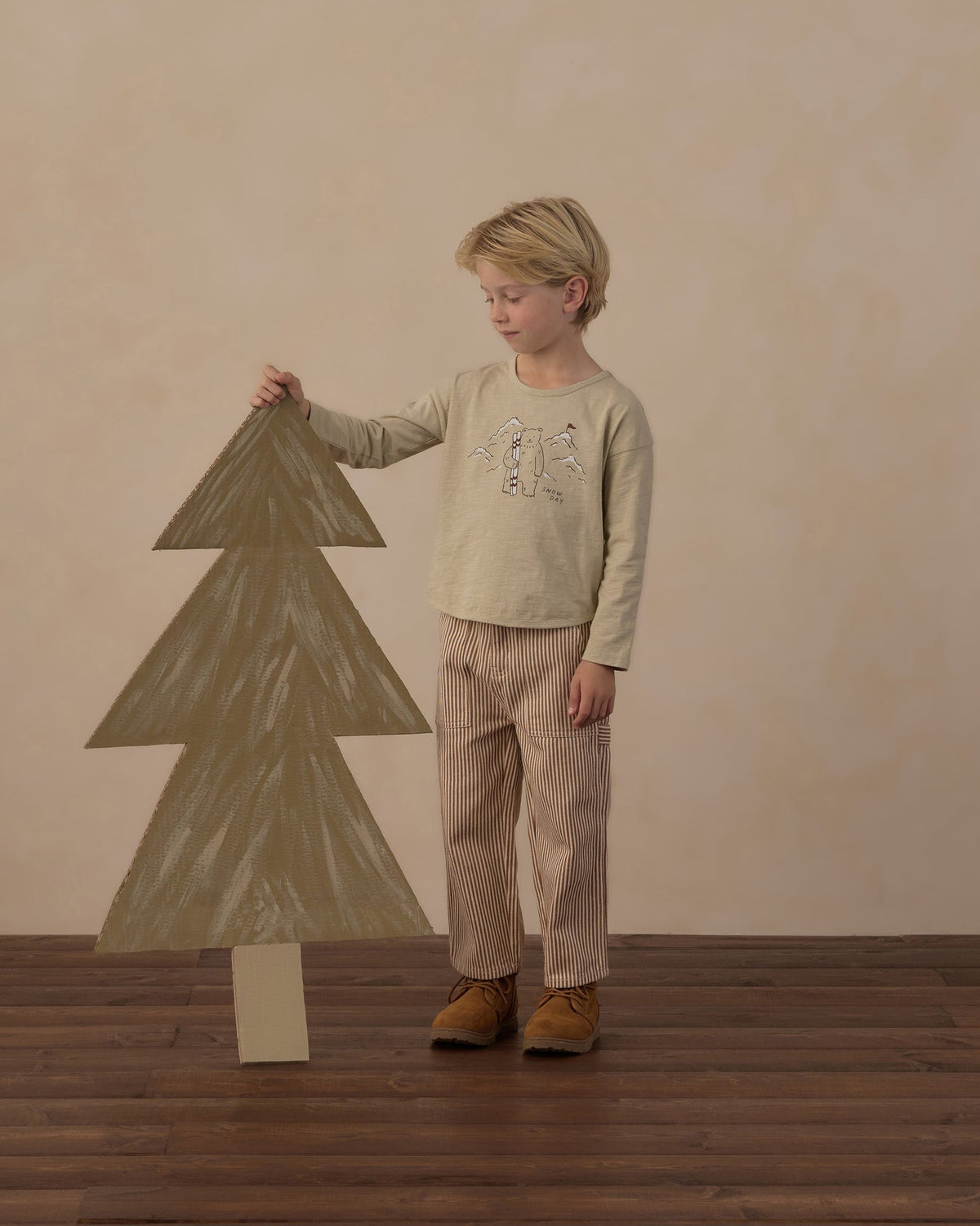 A young boy stands on a wooden floor against a textured beige wall, holding a large cardboard Christmas tree. He is wearing a light beige long-sleeve shirt with a graphic print and beige pinstriped pants.