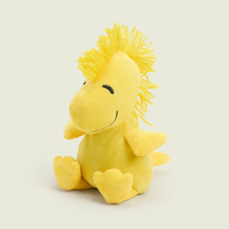 A bright yellow Woodstock Junior by Warmies plush toy sitting upright against a plain white background, featuring stitched closed eyes, soft outstretched wings, and a tuft of feathers on top of its head.