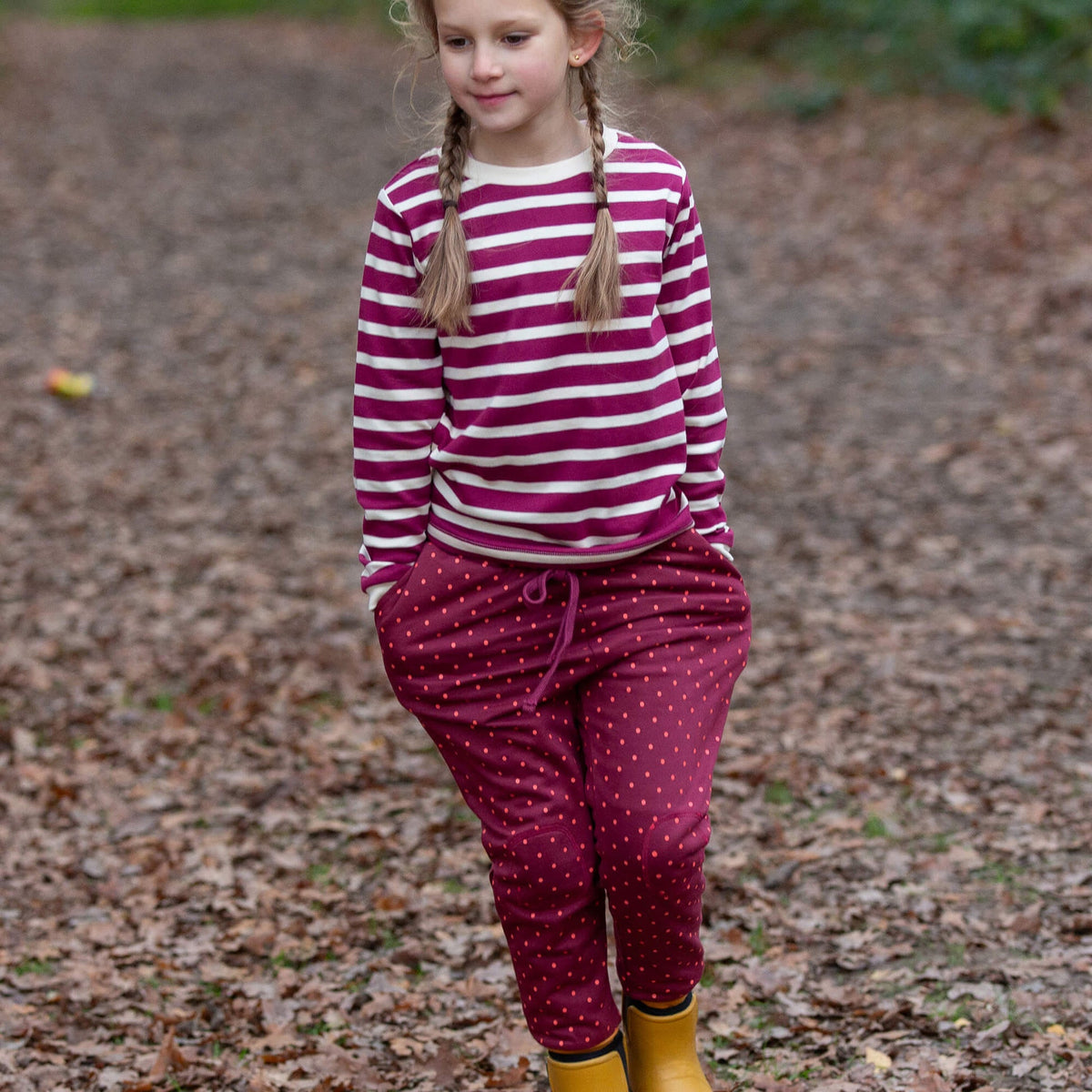 A young girl wearing maroon pants with red polka dots and yellow rain boots in a leafy outdoor setting