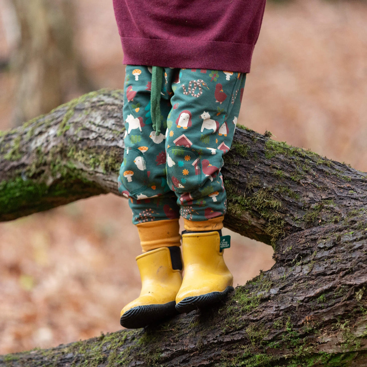 A child's legs wearing green patterned pants and bright yellow rain boots, standing on a mossy tree branch in an outdoor forest setting.