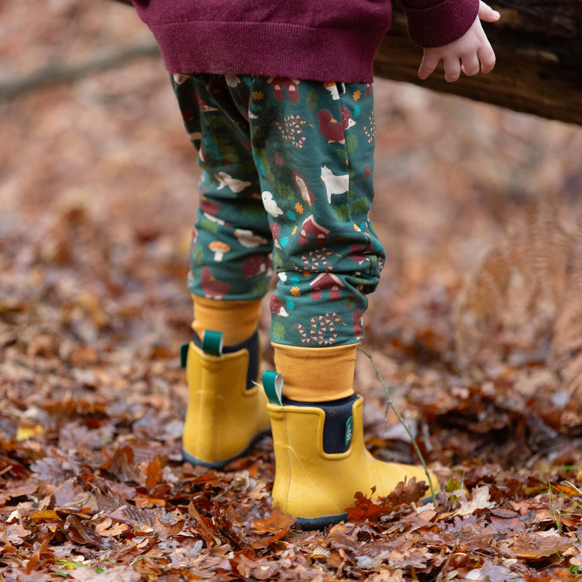 A child's legs wearing green patterned pants with autumn-themed prints, yellow rain boots, and black socks, standing on a ground covered in dry brown leaves.