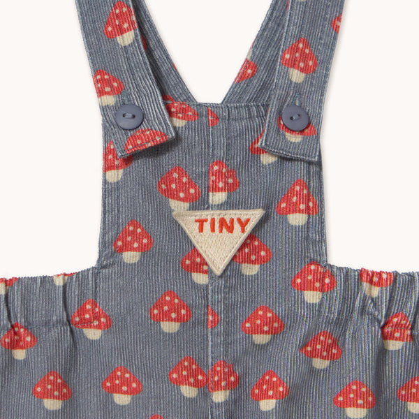 Close-up of a children's corduroy overall in a blue-gray color with a red and white mushroom print, featuring a triangular beige patch with the brand name 'TINY' on the chest.