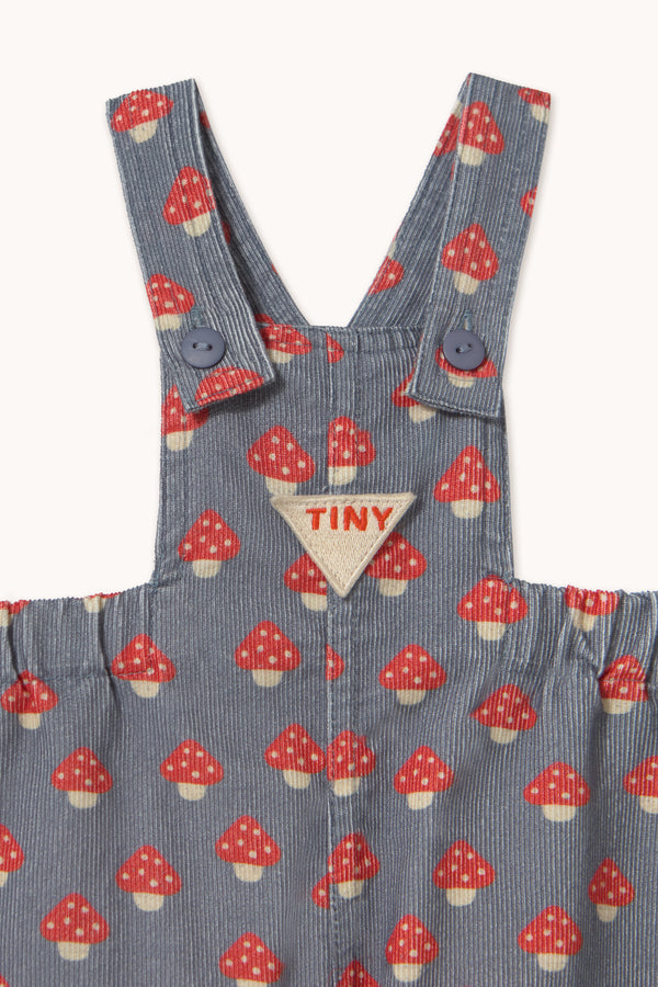 Close-up of a children's corduroy overall in a blue-gray color with a red and white mushroom print, featuring a triangular beige patch with the brand name 'TINY' on the chest.