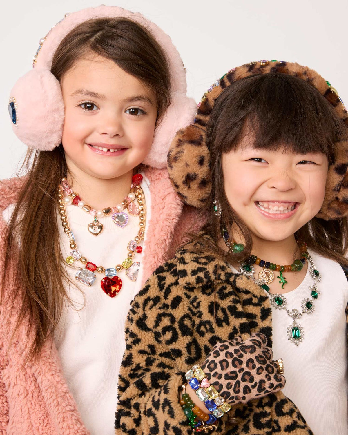 Two young girls smiling and posing together in cozy winter fashion. The girl on the left wears pink faux-fur earmuffs and a pink fuzzy cardigan over a white top, accessorized with colorful heart charm necklaces.