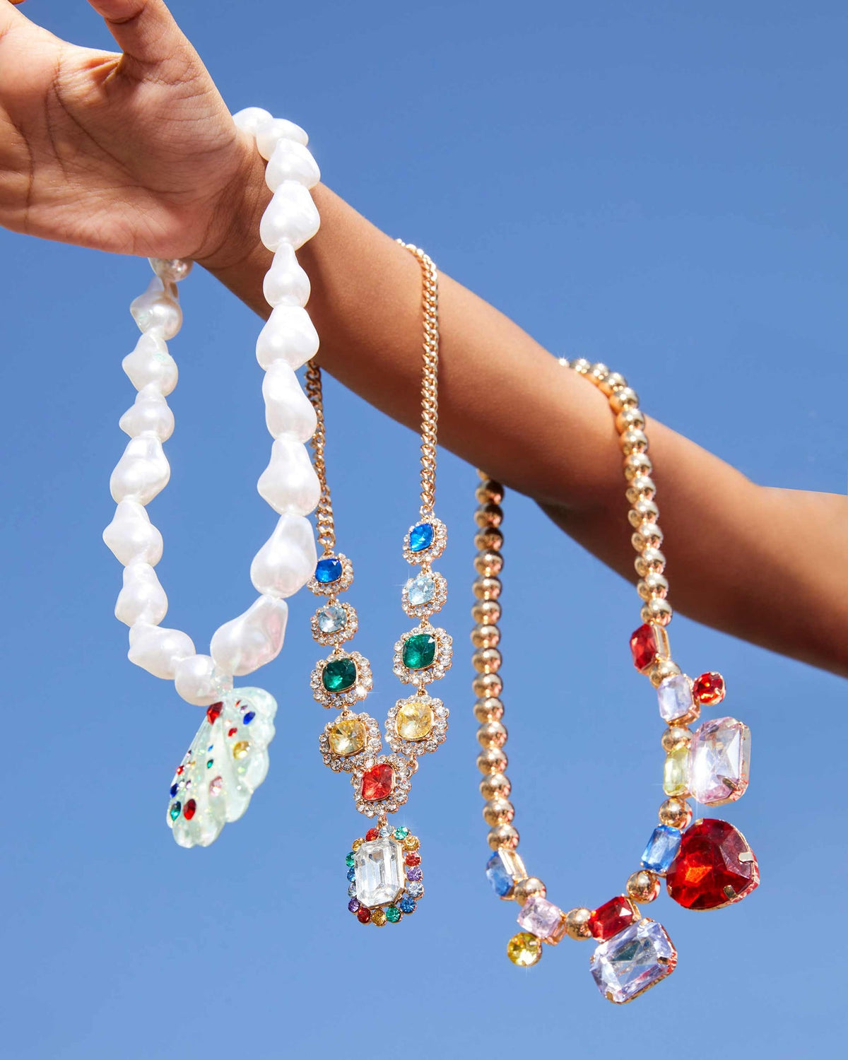 Three pearl necklaces with colorful gemstone pendants held against a blue sky.