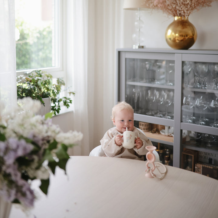 Baby holding a mug in a room with a window, plants, and a glass cabinet.