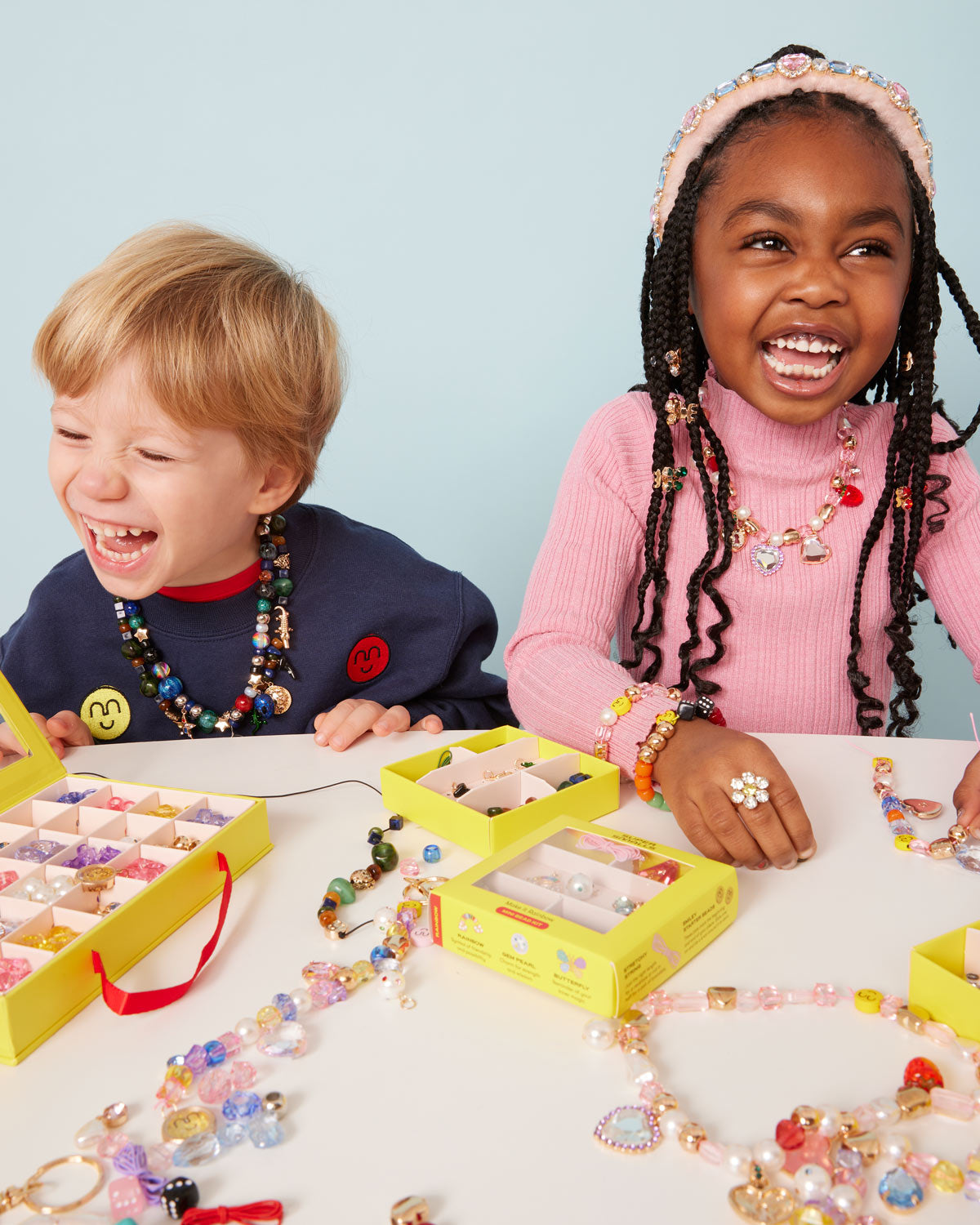 Two children smiling and laughing at a table filled with colorful handmade beaded jewelry and yellow display boxes, against a light blue background.
