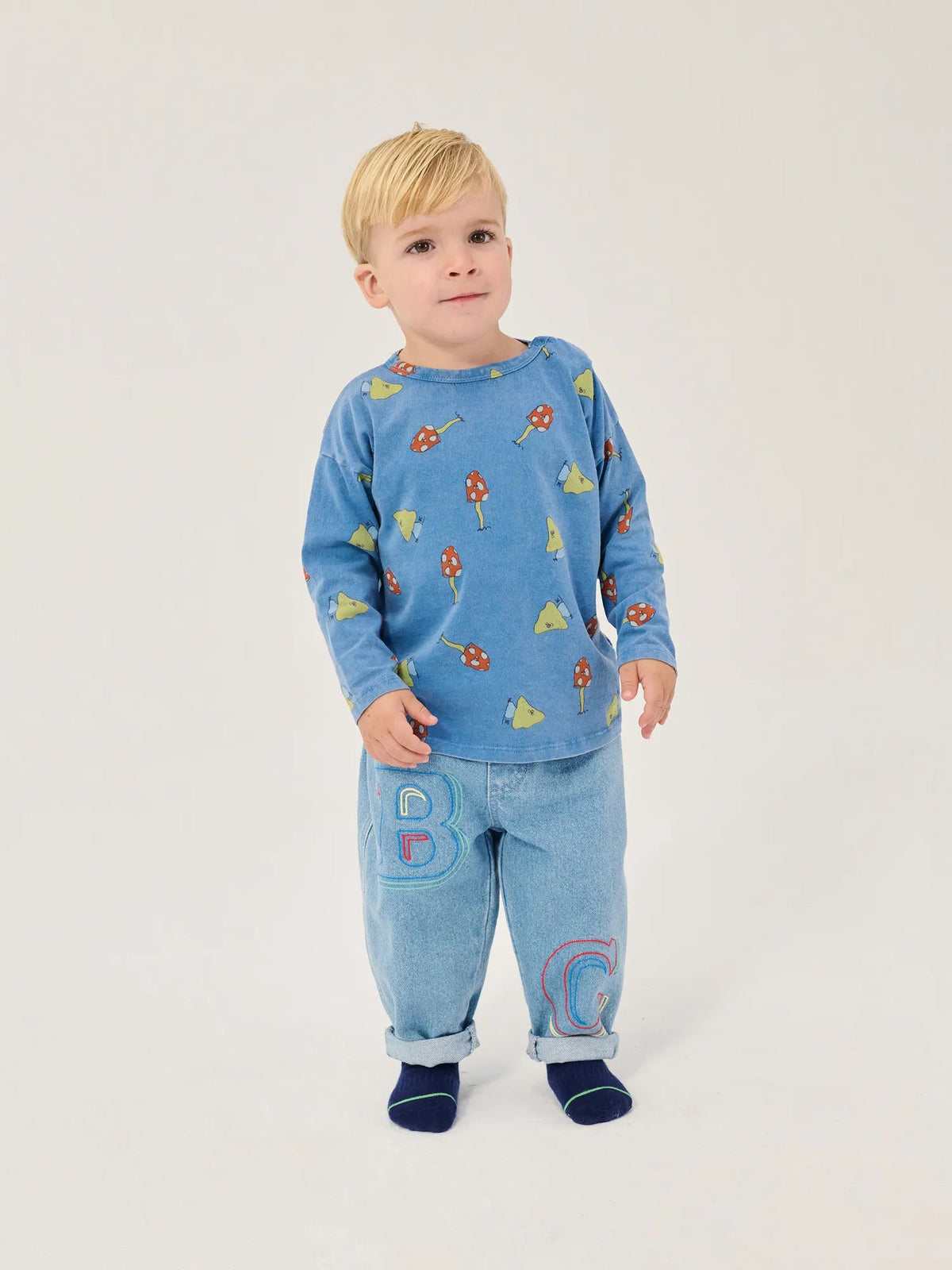 A young child wearing a light blue long-sleeve top with a colorful bug print and matching light blue pants with large embroidered letters, standing against a plain light background.
