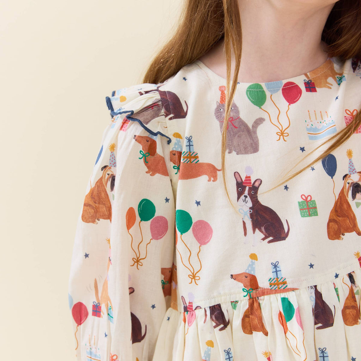 A close-up of a baby dress with a cream-colored base and an all-over print of dachshunds, birthday candles, party hats, balloons, and gift boxes