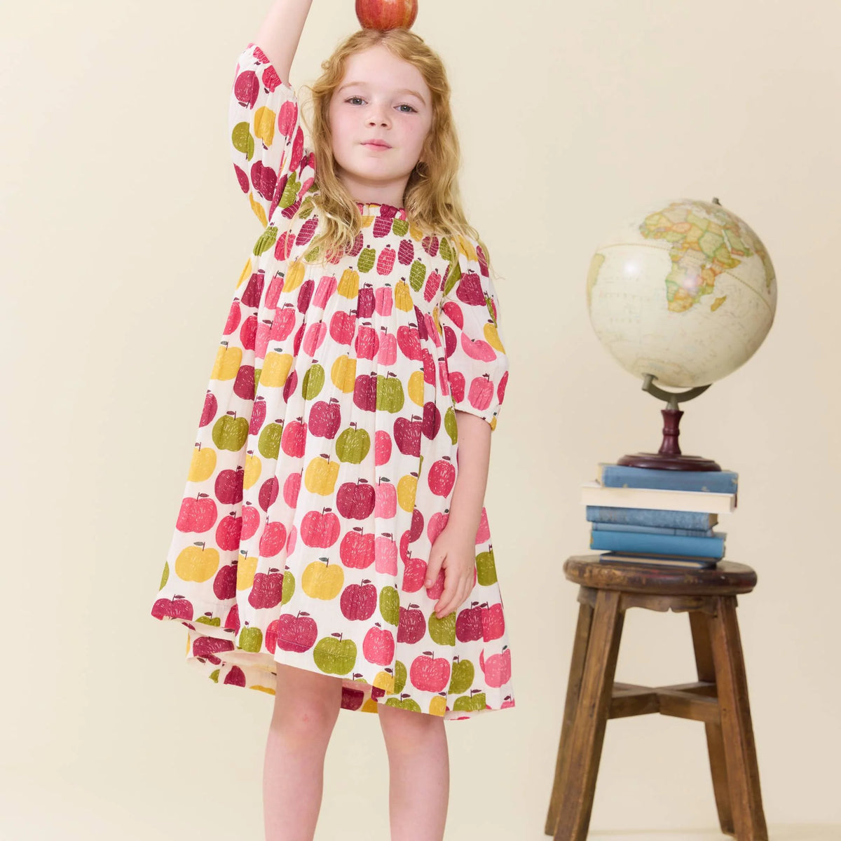 A young girl stands wearing a white short-sleeve dress covered in a colorful apple print. She is holding a red apple above her head, with a small wooden stool holding books and a globe beside her, against a plain cream background.
