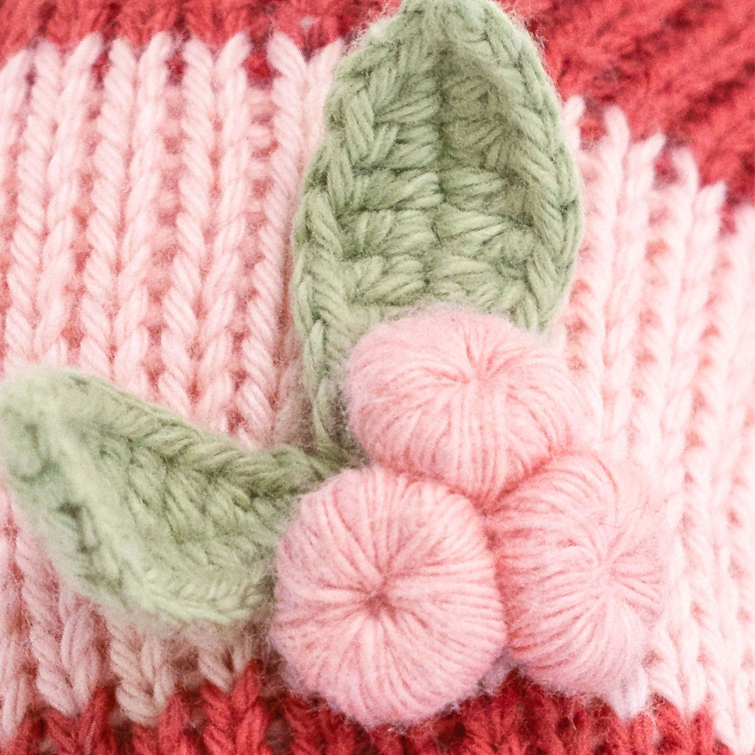 Close-up of a knitted textile featuring pink and coral-red stripes with a crocheted flower applique in light green and pink.