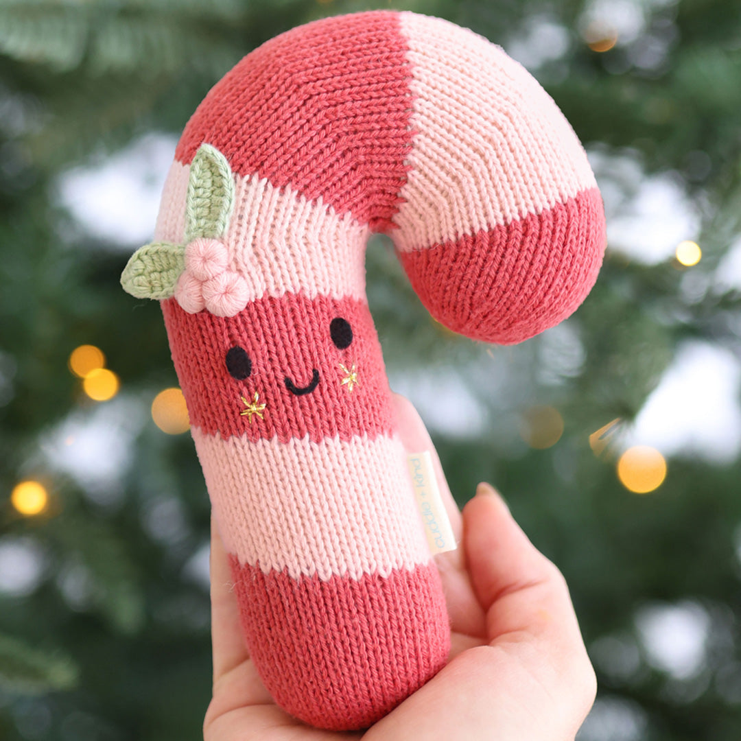 A hand holds a knitted candy cane plush toy with pink and white stripes, embroidered facial features, and a small green leaf and pink flower accent, against a blurred Christmas tree background with warm lights.