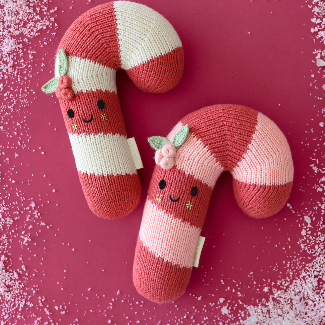 Two knitted candy cane pillows with smiling faces, decorated with small pink flowers and green leaves, set against a dark pink background with white faux snow.