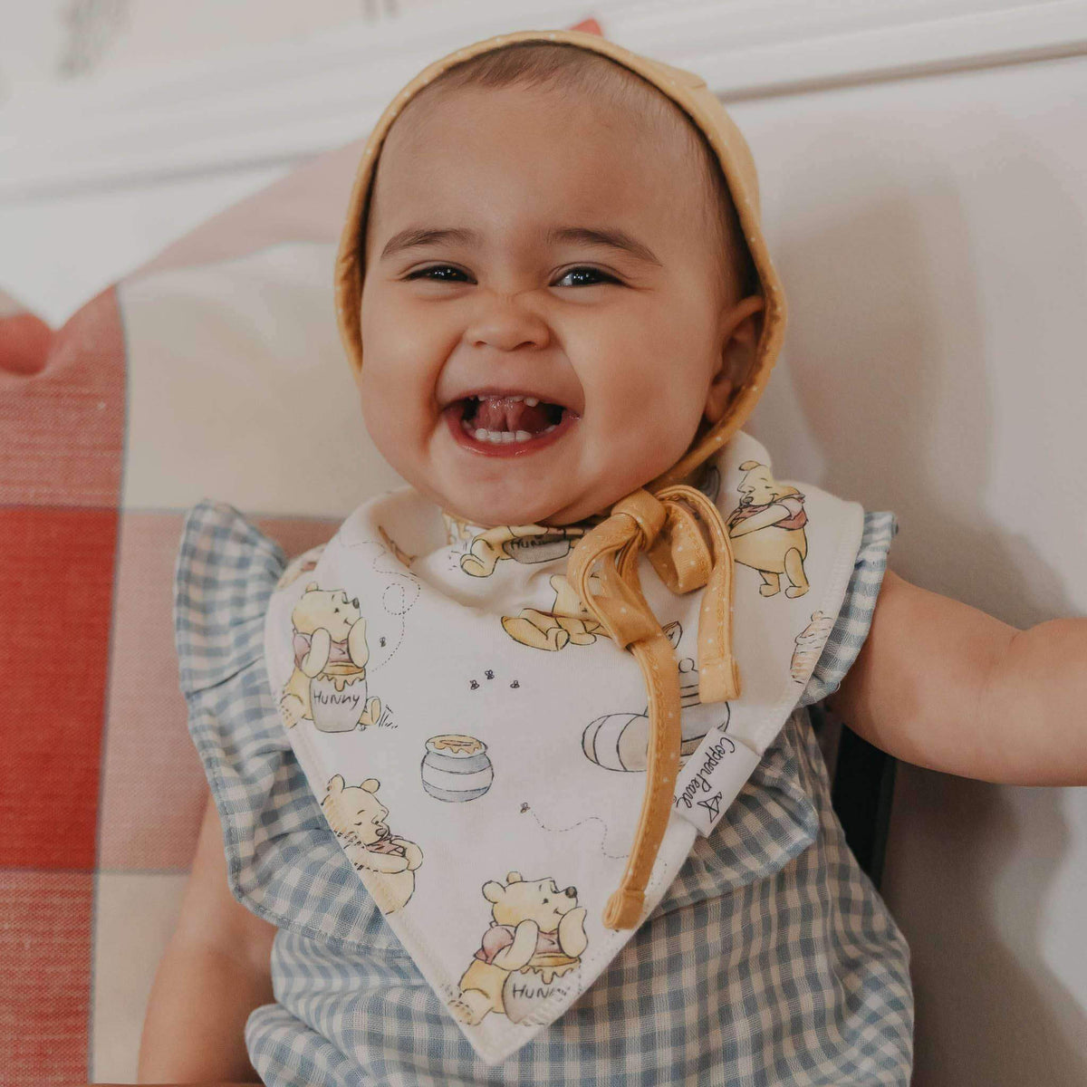 Baby wearing a Winnie the Pooh bib and bonnet, sitting on a couch.