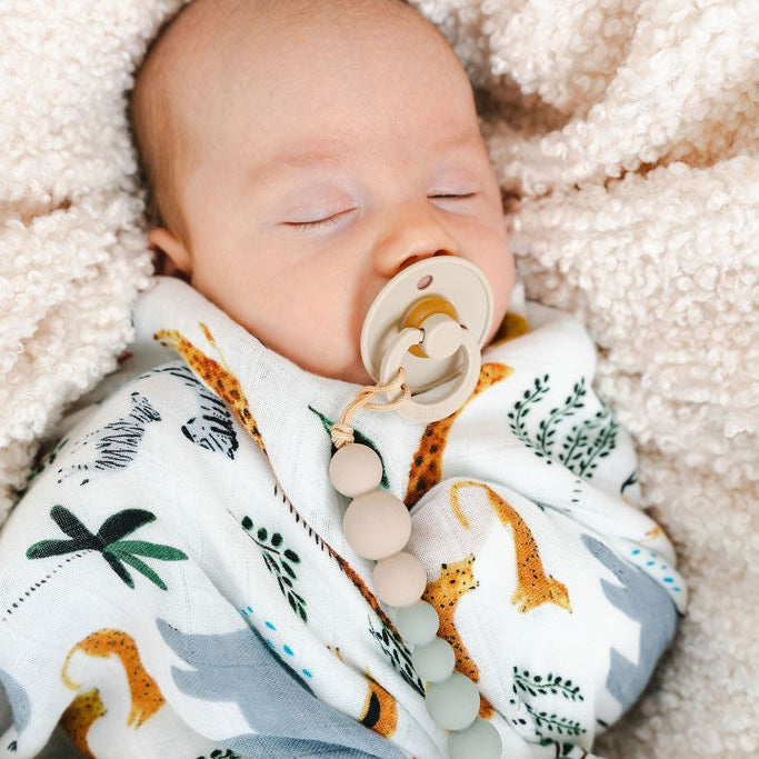 A sleeping newborn baby swaddled in a white blanket with a safari animal print, lying on a soft cream fleece blanket. The baby has a pacifier with wooden beads attached.