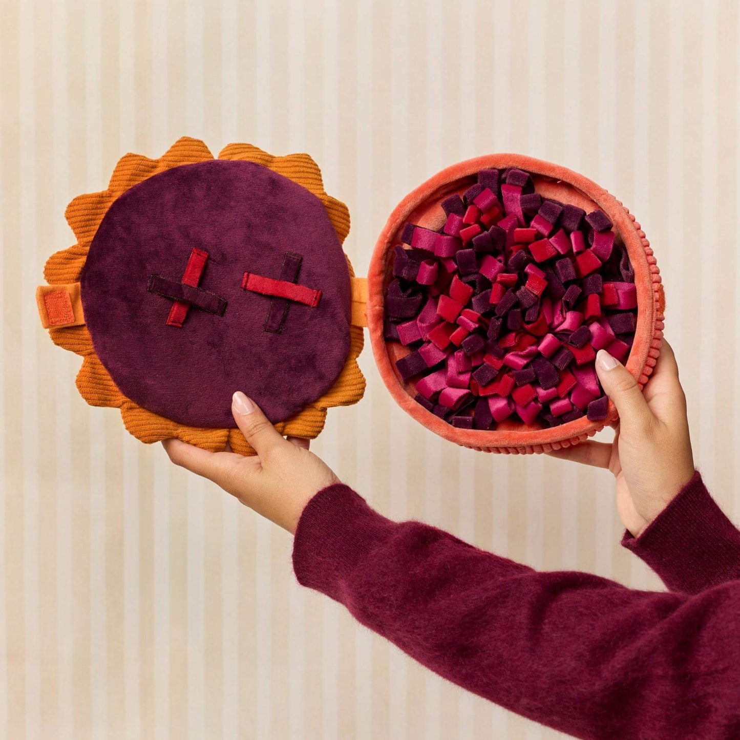 A person holds two round felt-like decorative pieces against a light beige striped background. One piece has a dark purple center with red and black ribbon crosses on an orange scalloped border.