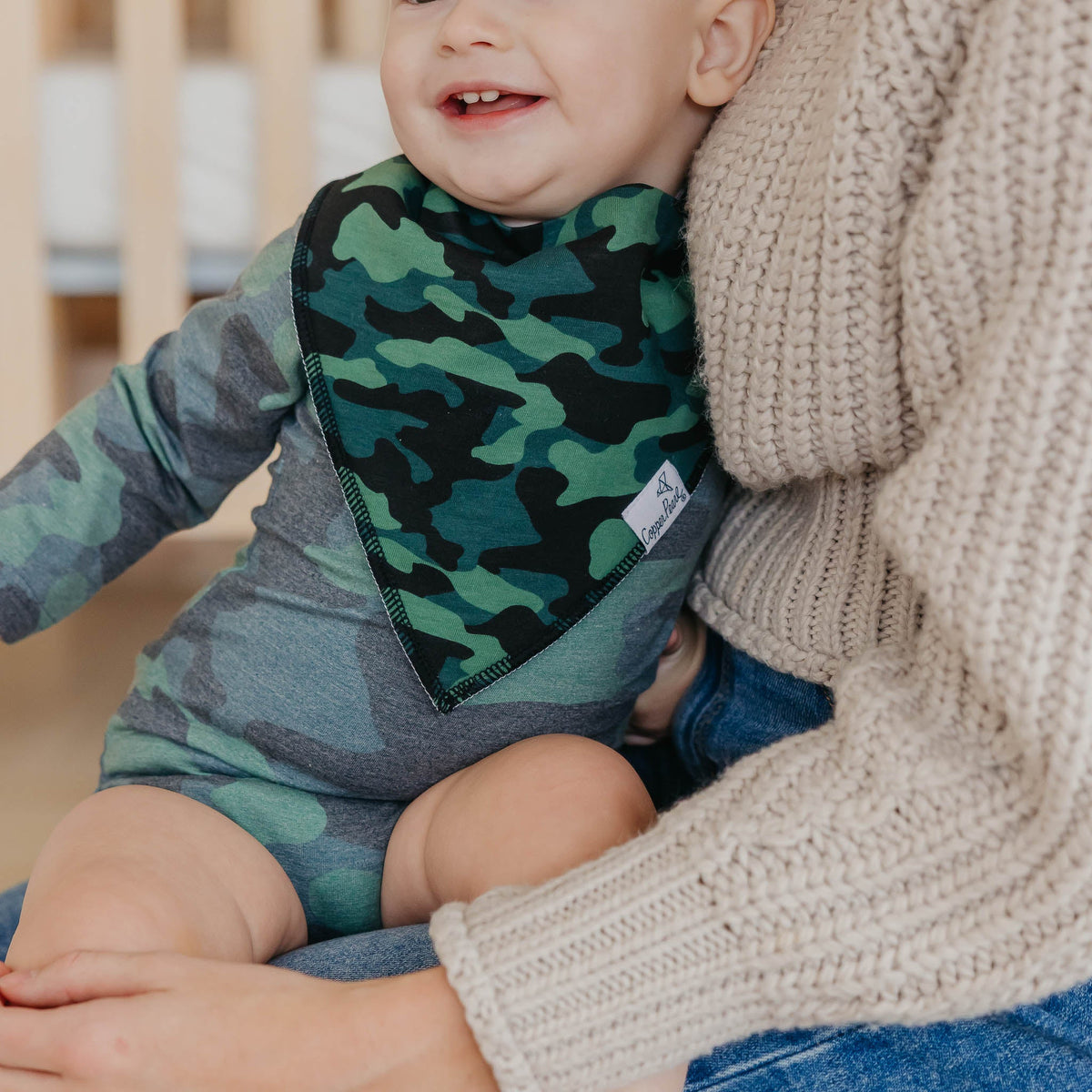 Child wearing a camouflage bib with a person holding them, sitting on a wooden floor.