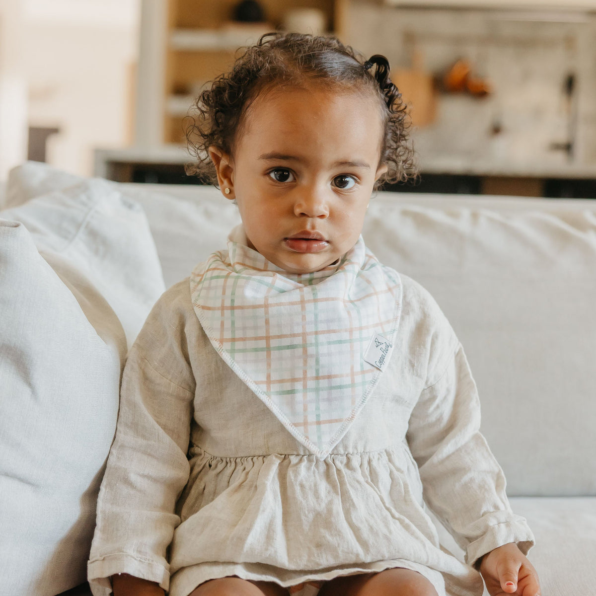 Child wearing a beige dress with a plaid bib in a kitchen setting