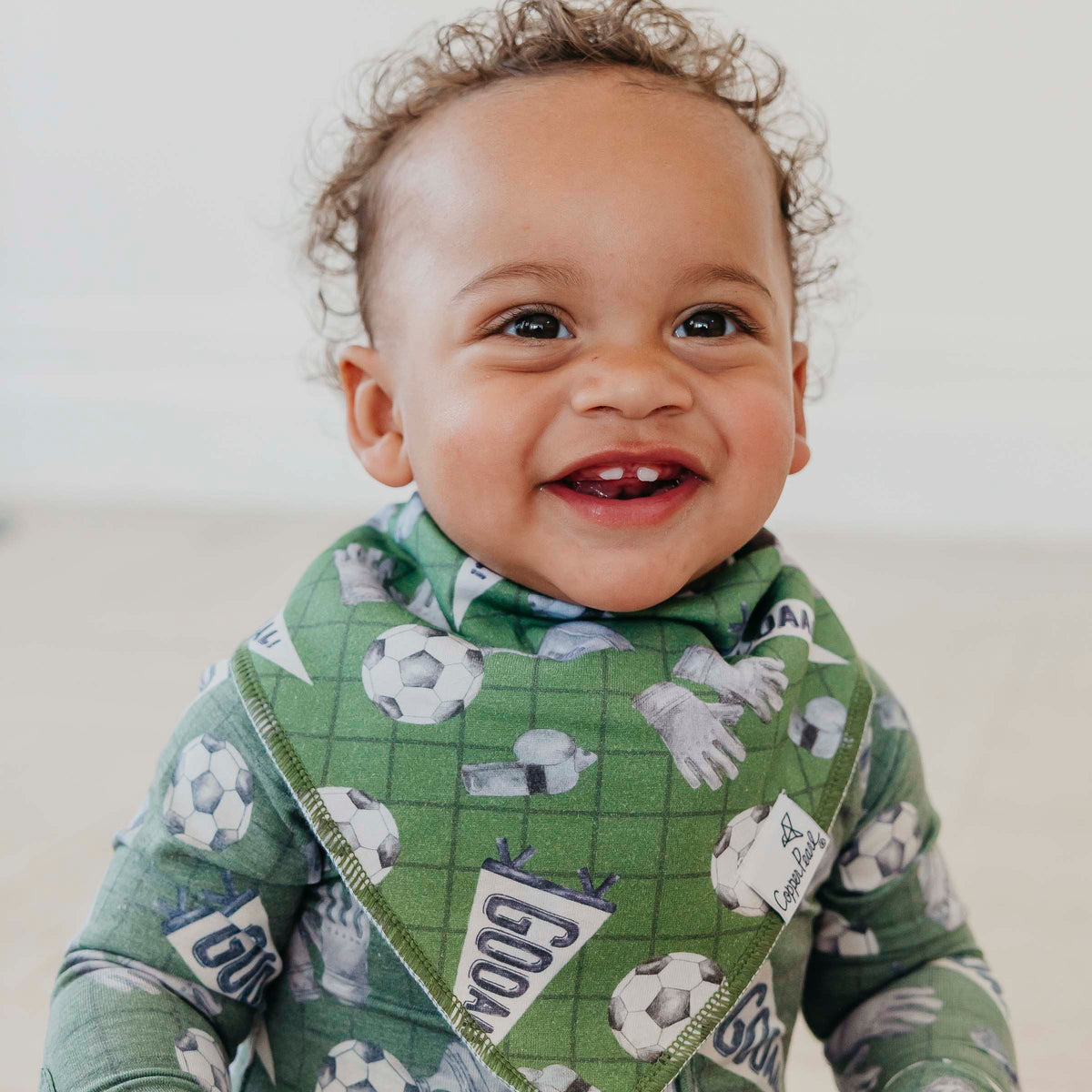 A smiling baby wearing a green bib and matching long-sleeve top with a soccer-themed print, photographed in a bright indoor setting.