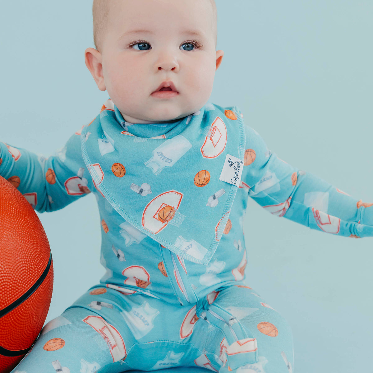 A baby wearing a light blue long-sleeve footless one-piece pajama with a basketball-themed print, sitting next to an orange basketball against a light blue background.