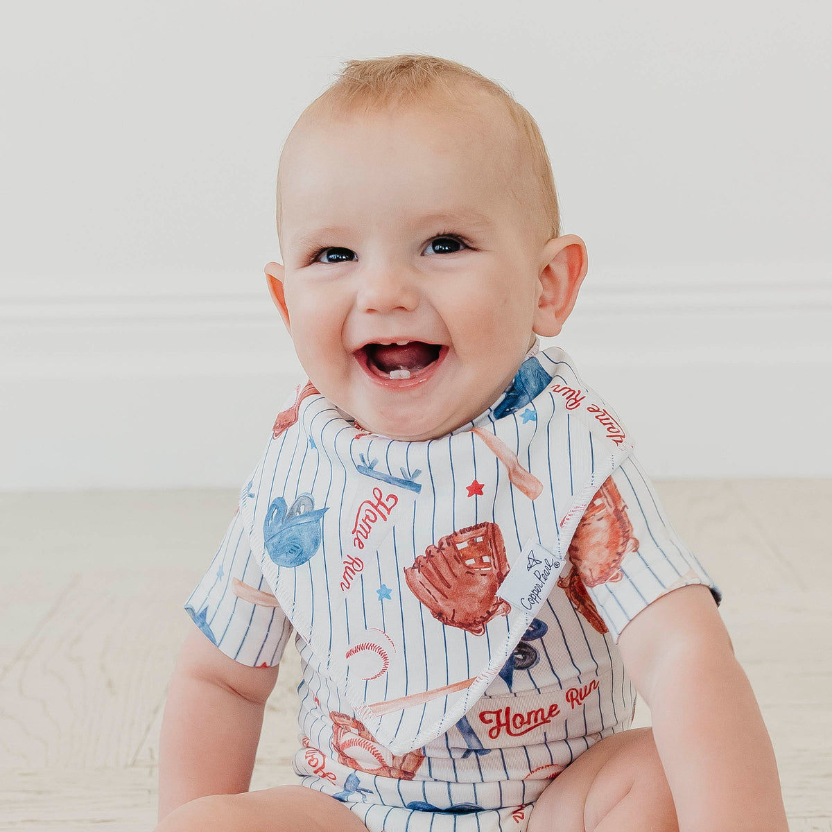 A smiling baby sitting on a light wood floor wearing a short-sleeve bib with a baseball pinstripe pattern and red, blue, and white graphics.