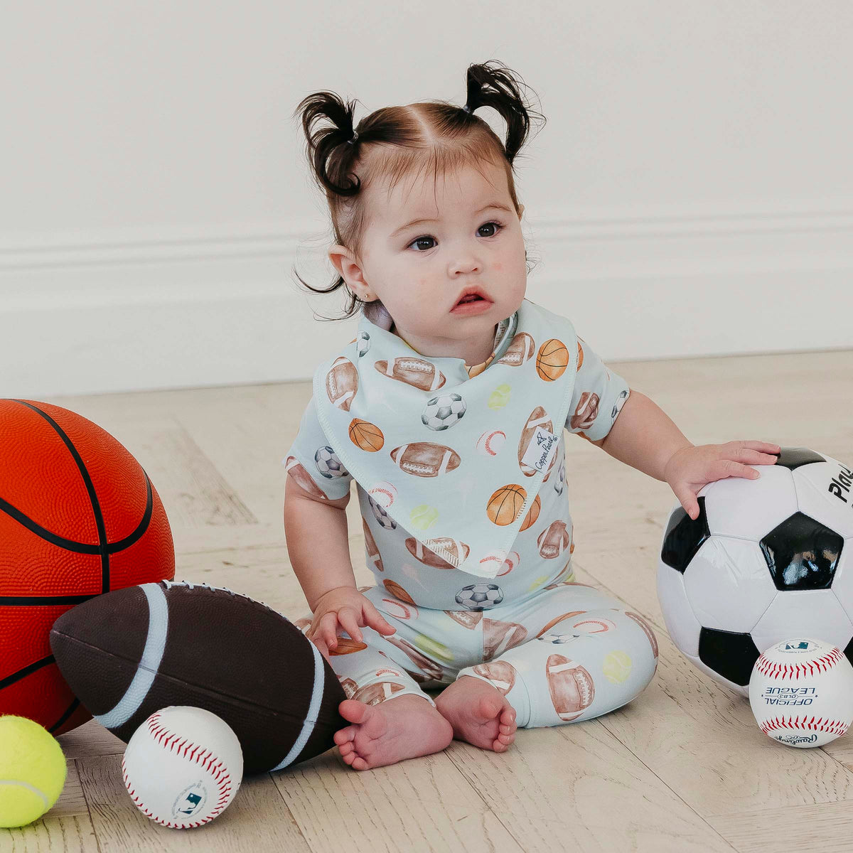 A baby sitting on a light wood floor surrounded by sports toys, wearing a light blue one-piece pajama set with a brown, beige, and white football pattern.