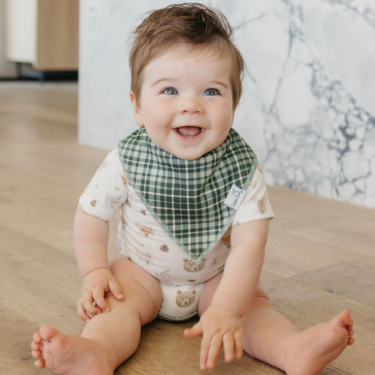 Baby sitting on the floor wearing a green knitted hat and white outfit with a pattern.