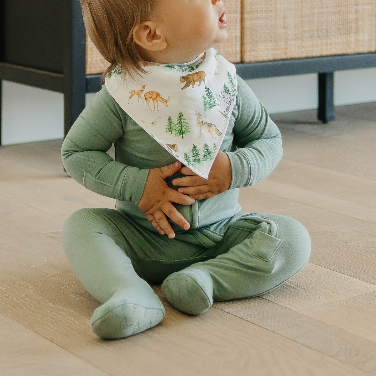 Baby in green outfit with forest-themed bib sitting on a wooden floor.