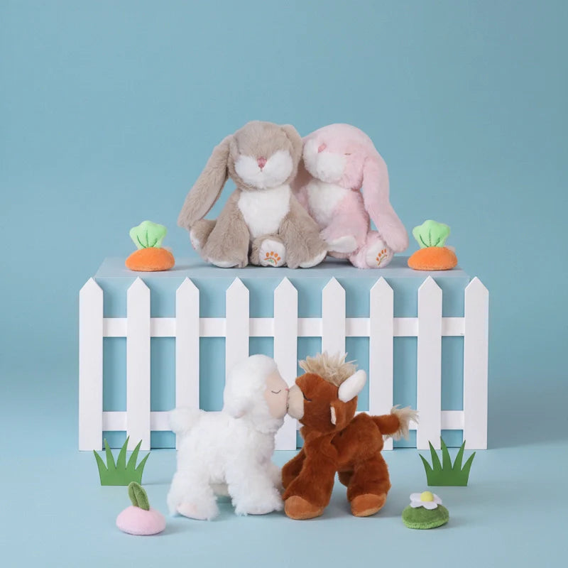 Plush toys including a gray bunny, pink rabbit, white lamb, and brown bear in front of a white picket fence on a blue background.