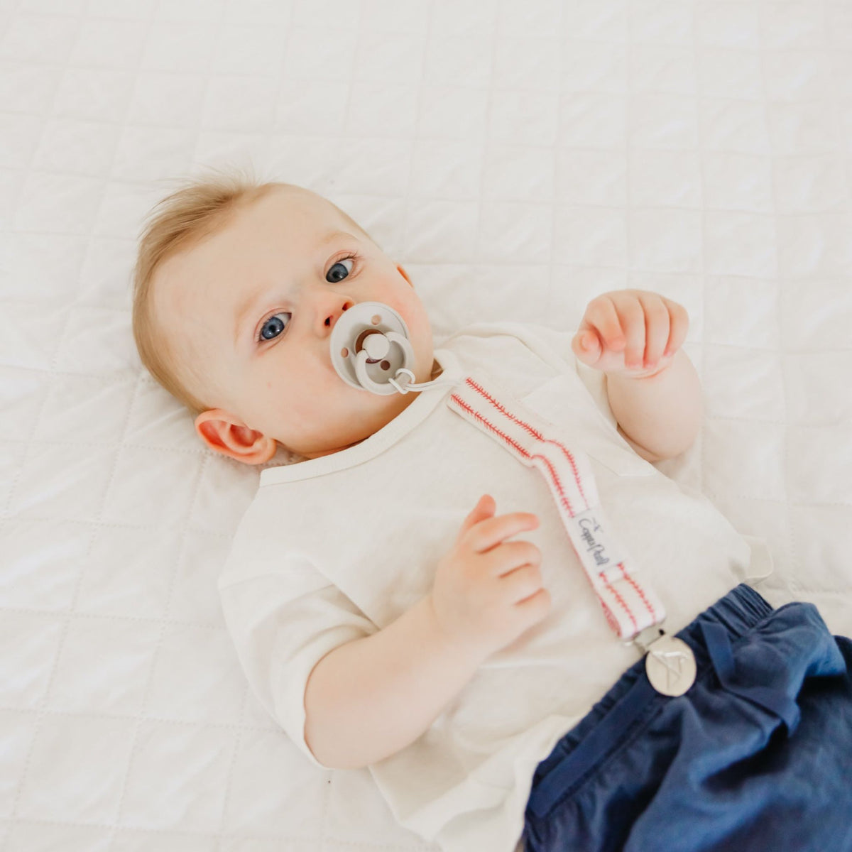 Baby lying on a white blanket wearing a white onesie with pink trim and blue pants.