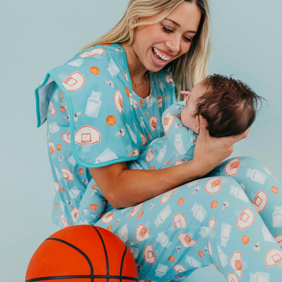 A woman and baby wearing matching light blue pajamas with a basketball-themed print, with the woman holding the baby. An orange basketball is visible in the foreground against a solid light blue background.