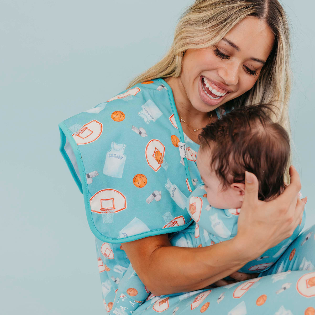 A smiling woman holds a baby wearing matching light blue sleepwear with an orange and white basketball-themed print, against a solid light blue background.