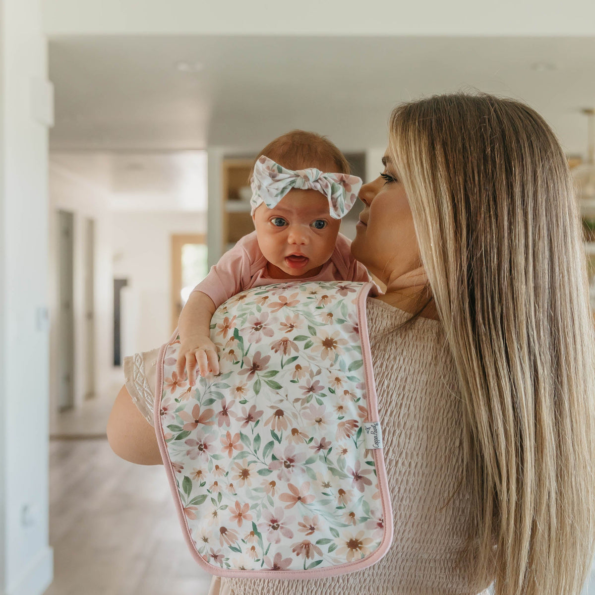 Woman holding a baby wrapped in a floral blanket inside a home.