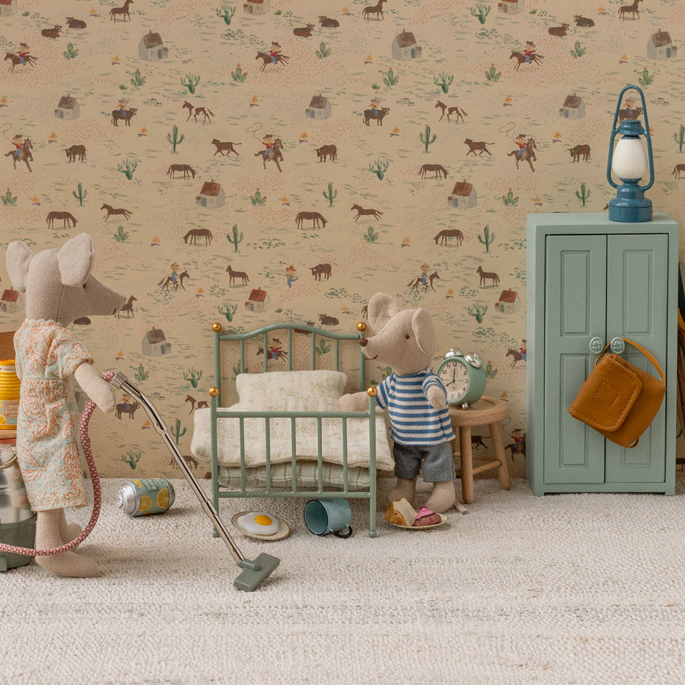 Two Maileg mouse toys in a miniature room setting with patterned wallpaper and carpet; one mouse on the left operates a toy vacuum cleaner next to a thermos and canister, while another mouse on the right stands beside a mint green single bed and a matching storage cabinet with a blue lantern on top.