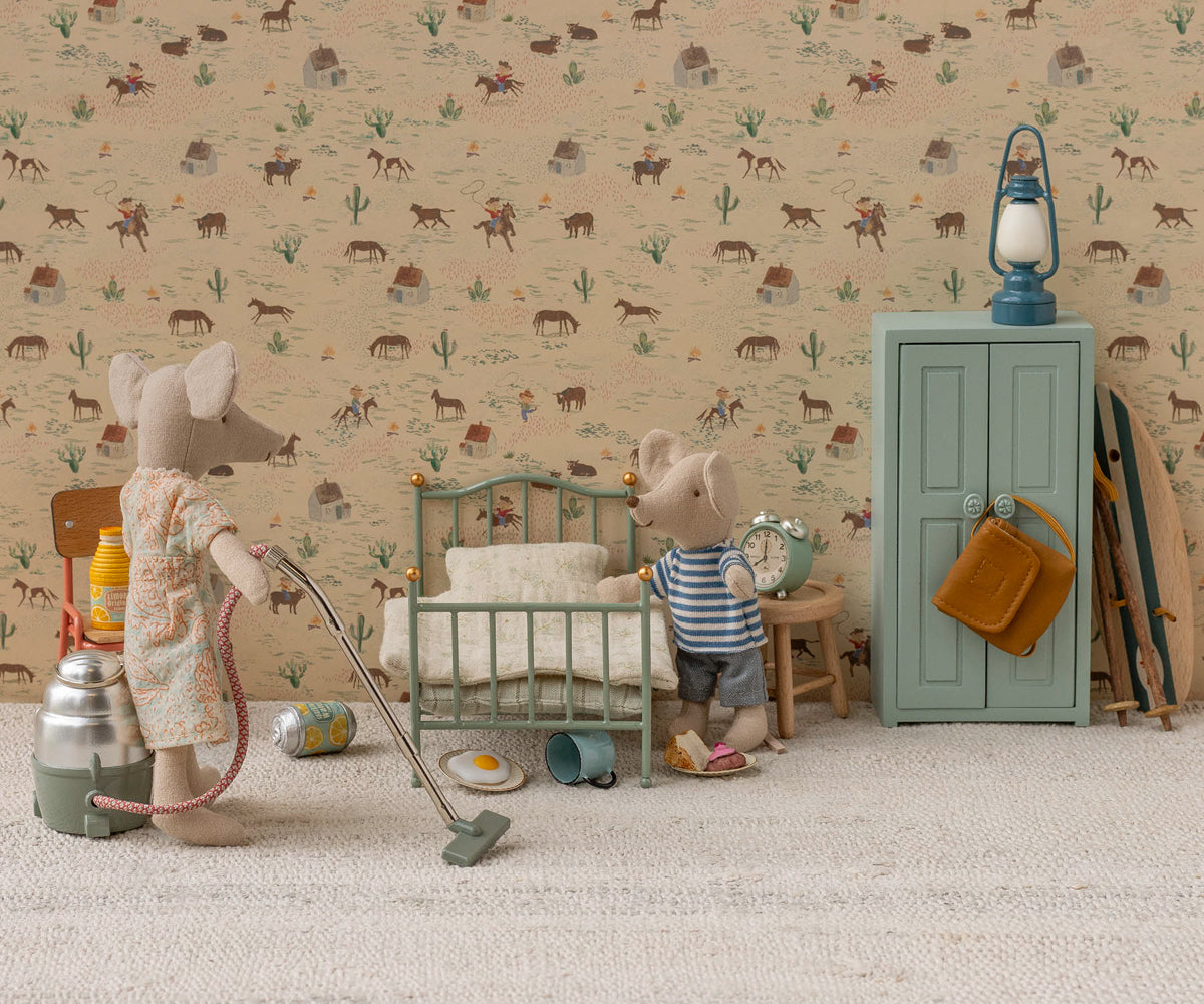 Two Maileg mouse toys in a miniature room setting with patterned wallpaper and carpet; one mouse on the left operates a toy vacuum cleaner next to a thermos and canister, while another mouse on the right stands beside a mint green single bed and a matching storage cabinet with a blue lantern on top.
