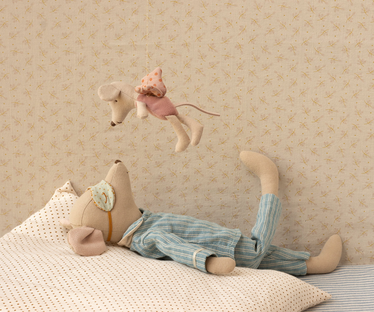 Stuffed animal lying on a bed with another animal hanging above, against a patterned wall.