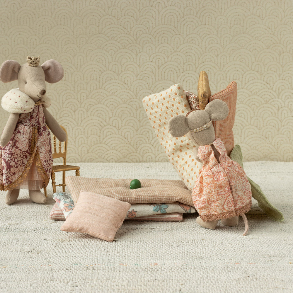 A Maileg toy set showing a Big Sister mouse standing next to a stack of patterned mattresses, a miniature pillow, and a tiny green pea resting on the bed setup.