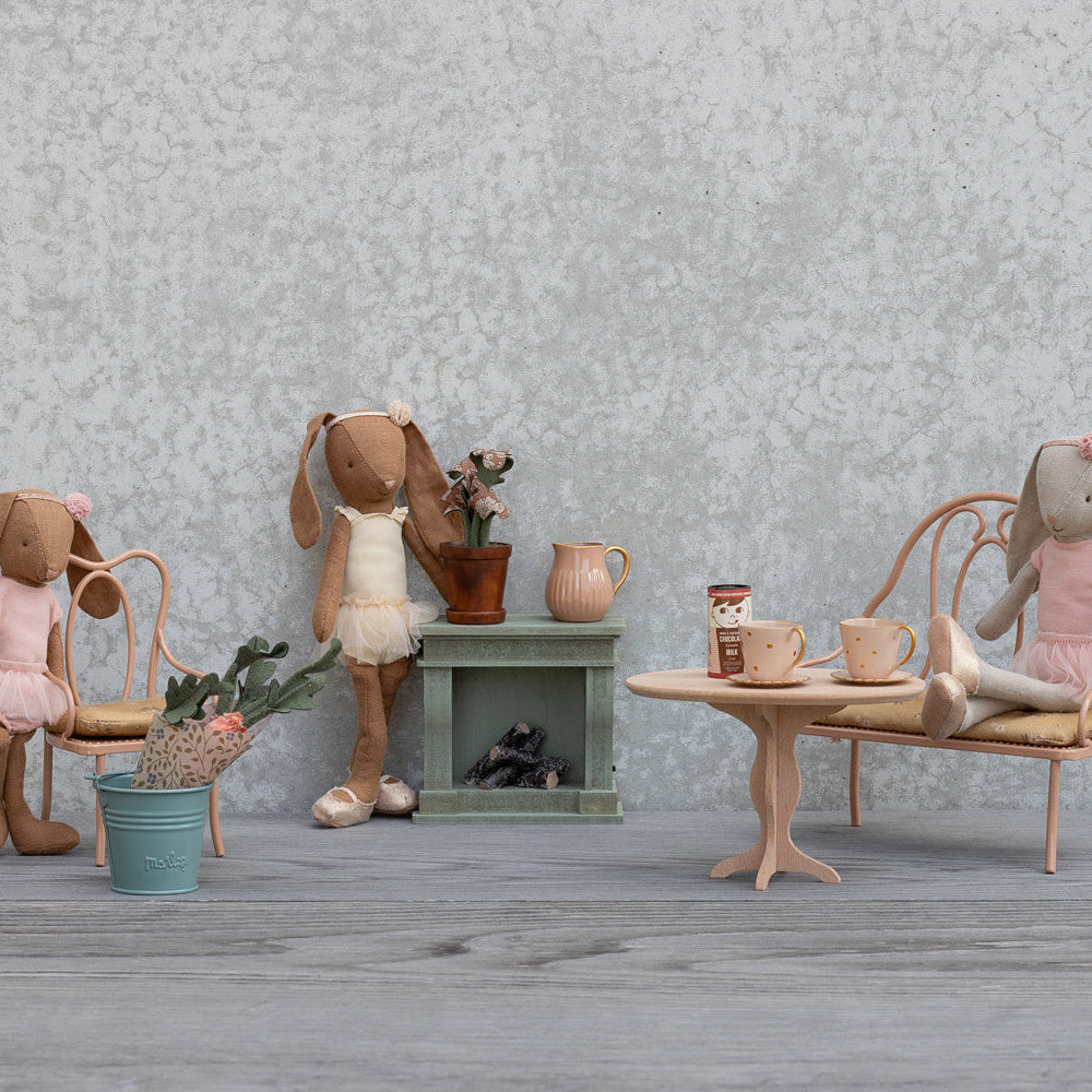 Two Maileg toy rabbits are sitting in a miniature room with a table, chairs, a small fireplace, and decorative accessories including potted plants and a crate of carrots.